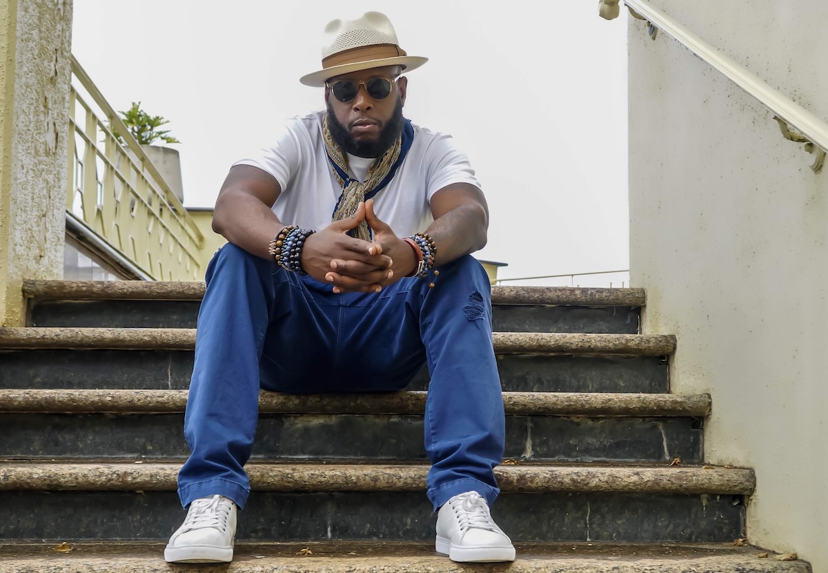 Rapper Talib Kweli sits on outdoor stairs wearing a fedora, sunglasses, white t-shirt, blue pants, and white sneakers with hands clasped.